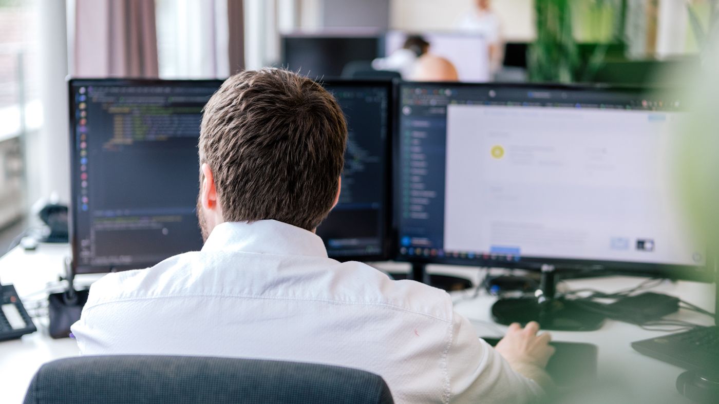 TYPO3-Mitarbeiter bei der Arbeit Junger Mann sitzt vor zwei Bildschirmen in einem modernen Büro, schulterblick Fotografie