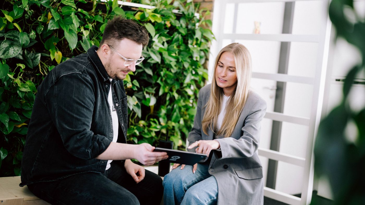 Zwei Personen sitzen in einem modernen Büro mit vielen Pflanzen und schauen gemeinsam auf ein Tablet