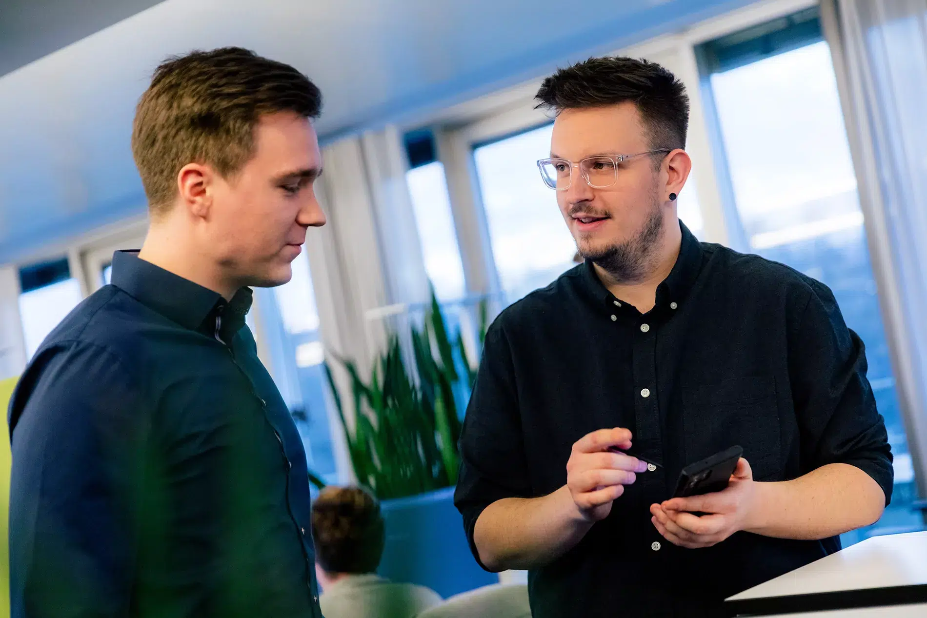 TYPO3-Agentur in Nürnberg – Experten im Gespräch Zwei Personen führen in einem Büro ein Gespräch. Einer hält ein Telefon und gestikuliert, während der andere zuhört. Im Hintergrund sind Zimmerpflanzen und große Fenster zu sehen.